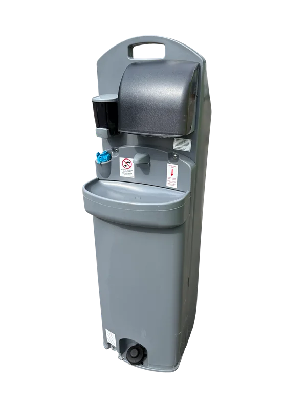 A tall, slender, grey standalone portable handwashing station. It features a foot pump at the base for hands-free operation, a central sink basin with a blue spigot, a dark grey paper towel dispenser near the top, and a small black soap dispenser mounted to the left side